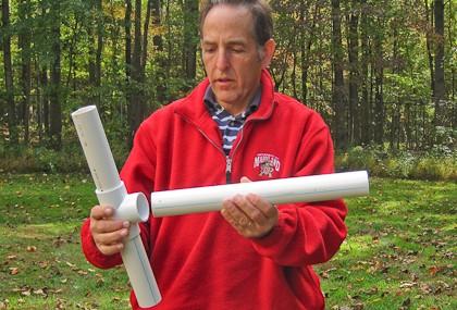 A person inserts a piece of PVC pipe into the center socket of a T-shaped joint that already has two shorter pipe pieces inserted into the side sockets.