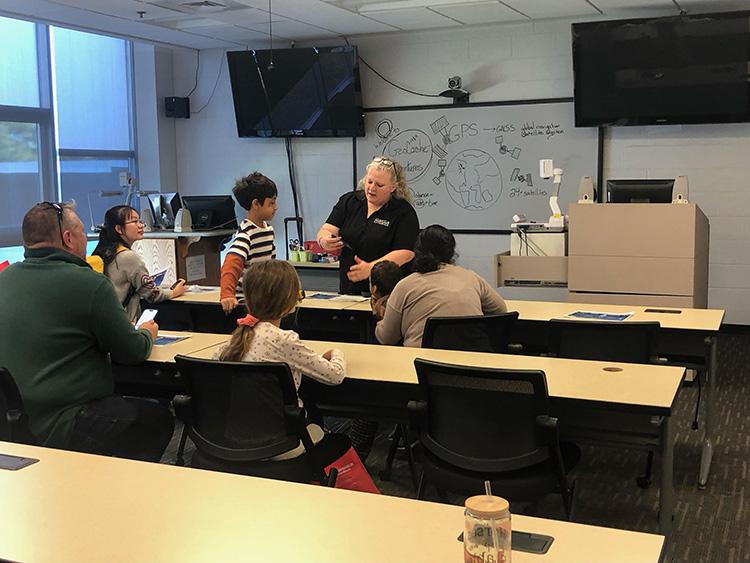 A woman is explaining something to a small group of adults and children in a classroom. A whiteboard with diagrams is in the background.