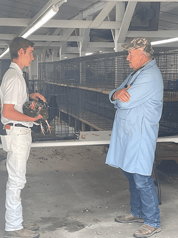  A young man in a white shirt and pants holds a chicken, talking to an older man in a blue coat and camo hat. They're in a barn with cage-lined walls.