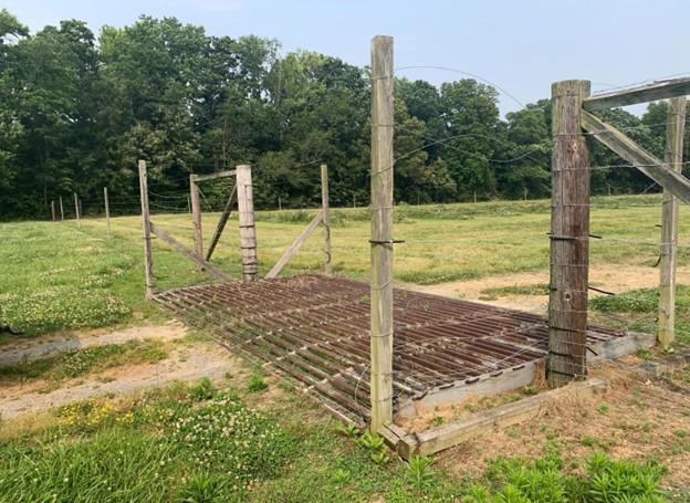 A metal cattle guard installed in an opening of a fenced pasture. The guard consists of parallel steel bars set over a shallow pit, creating a surface that vehicles can drive over but animals avoid. Wooden fence posts and wire fencing border both sides of the opening, with angled wooden braces supporting the structure. Beyond the cattle guard is an open grassy field, and a line of dense trees forms the background under a hazy sky.