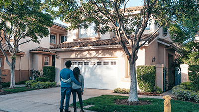 A couple stands arm in arm, facing a two-story stucco house with a tile roof and a white garage door. Green trees and shrubs surround the home, creating a hopeful, serene ambiance.