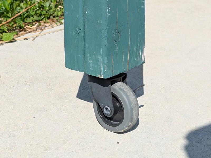 Close-up view of the base of a two-piece Salad Table leg, showing the board ends flush with each other. A lockable wheel is attached to the base of the leg.