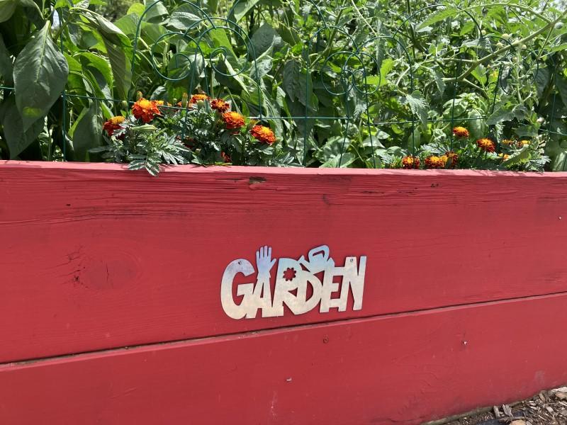 Close-up of the side of a wooden raised bed sealed with red paint and decorated with a metal “garden” sign attached to a board.