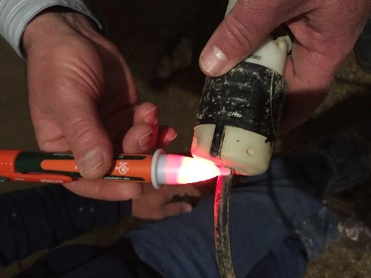 Close-up of two hands using a glowing red voltage tester on an exposed electrical wire connected to a white-and-black fitting. The tester tip illuminates to indicate live voltage, while the wire and connection are examined in a dimly lit indoor space.