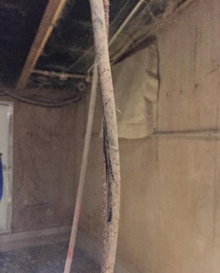 Dust-covered electrical cable hanging vertically in an unfinished basement or crawlspace. The outer cable jacket is split open along one side, exposing the inner conductors. Wooden framing, insulation, and unfinished wall surfaces are visible in the background under dim lighting.