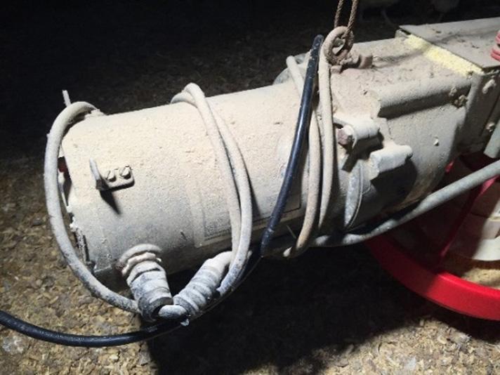 Dust-covered electric motor in an agricultural or industrial setting with several power cords and cables wrapped around the motor housing. A black cord and light-colored cables are looped multiple times around the cylindrical motor body, with additional wiring connected at one end. The motor is mounted above a red piece of equipment, and the surrounding area is dimly lit with dust and debris visible on nearby surfaces.
