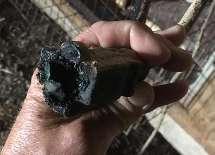 Close-up of a damaged electrical connector held in a person's hand. The end of the connector is heavily charred and melted, with black soot, corrosion, and heat damage visible around the opening. A dusty electrical cord extends from the back of the connector, and a wire mesh enclosure and wooden surfaces are visible in the background.