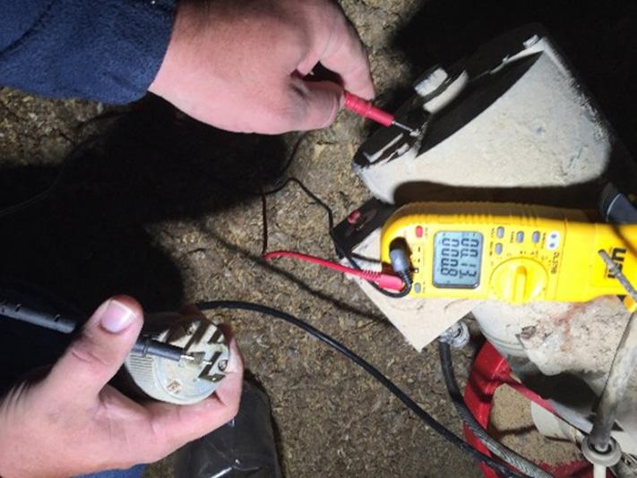 Close-up of electrical testing being performed on equipment in a dusty agricultural or industrial setting. Two hands hold multimeter probes against electrical terminals on a motor housing while a yellow digital multimeter displays a measurement. Electrical cords, wiring, and a red piece of machinery are visible around the motor, which is coated with dust and debris.