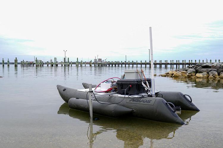 Autonomous surface vehicle (ASV) designed for aquatic research floating in shallow water near a shoreline. The small pontoon-style vessel is equipped with onboard sensors, electronics, and antennas used to collect oyster reef and environmental data. A pier, pilings, and rock breakwater are visible in the background.