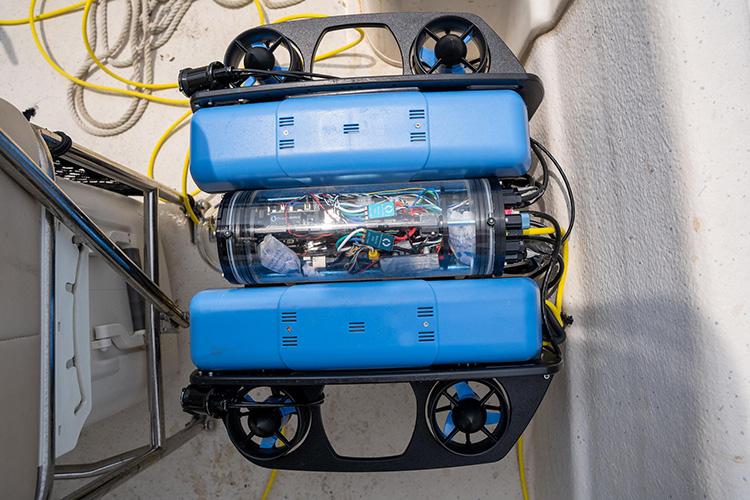 Blue ROV remotely operated underwater vehicle resting on a boat deck. The compact research vehicle is equipped with four visible propellers, onboard cameras, sensors, and electronics housed within a transparent pressure-resistant chamber. A tether cable connects the vehicle to surface controls, allowing it to collect underwater imagery and environmental data for oyster reef monitoring and research.