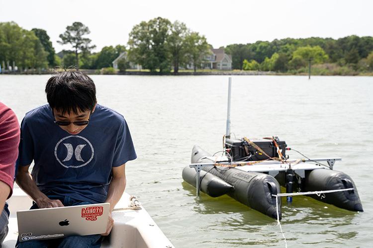 Student uses a laptop to communicate with and control an autonomous surface vehicle (ASV) floating nearby in a waterway. The pontoon-style research vessel is equipped with onboard electronics, sensors, and antennas used for oyster reef monitoring and environmental data collection. The image illustrates the use of computer-guided technology to operate and monitor the ASV during field research.