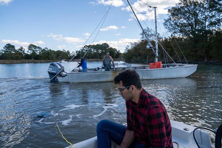 : A student operates a remotely operated underwater vehicle (ROV) from a small boat while conducting field research near an oyster harvesting vessel. The tethered ROV floats at the water's surface beside the boat, connected by a yellow cable. In the background, crew members work aboard a commercial oyster harvesting vessel anchored in a calm waterway bordered by trees. The image illustrates the use of underwater robotics to collect data in active oyster harvesting areas.