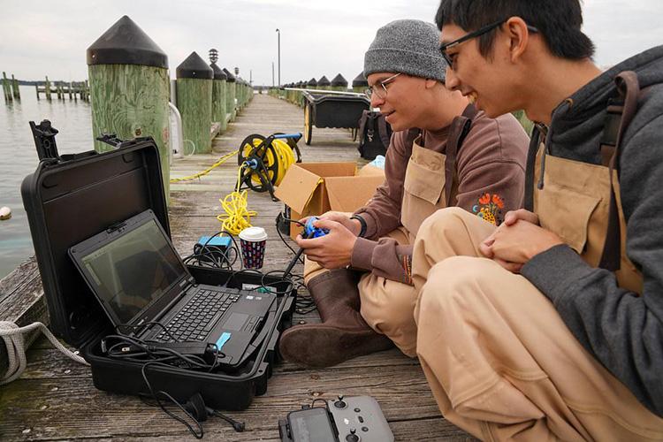 : Two students sit on a pier operating a remotely operated vehicle (ROV) for underwater research and data collection. One student uses a handheld controller while both view the live video feed displayed on a laptop housed in a protective case. Cables connecting the ROV extend across the dock toward the water, with pilings and a marina visible in the background.