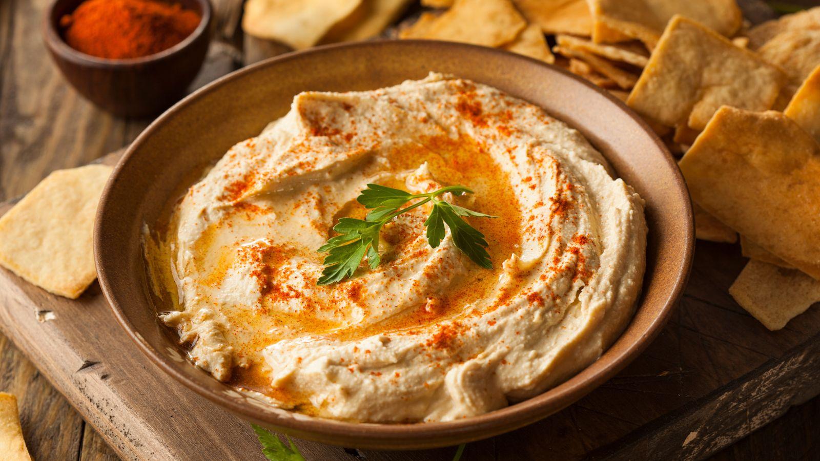 Bowl of creamy hummus sitting on cutting board with pita chips next to it and a spice bowl
