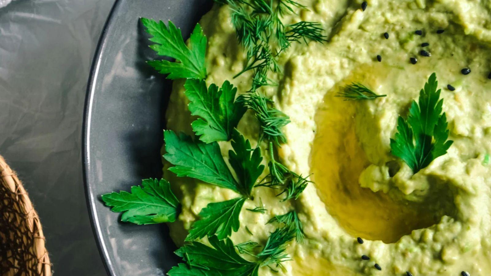 bowl of lemon kale hummus with herbs on top sitting on a plate next to a basket