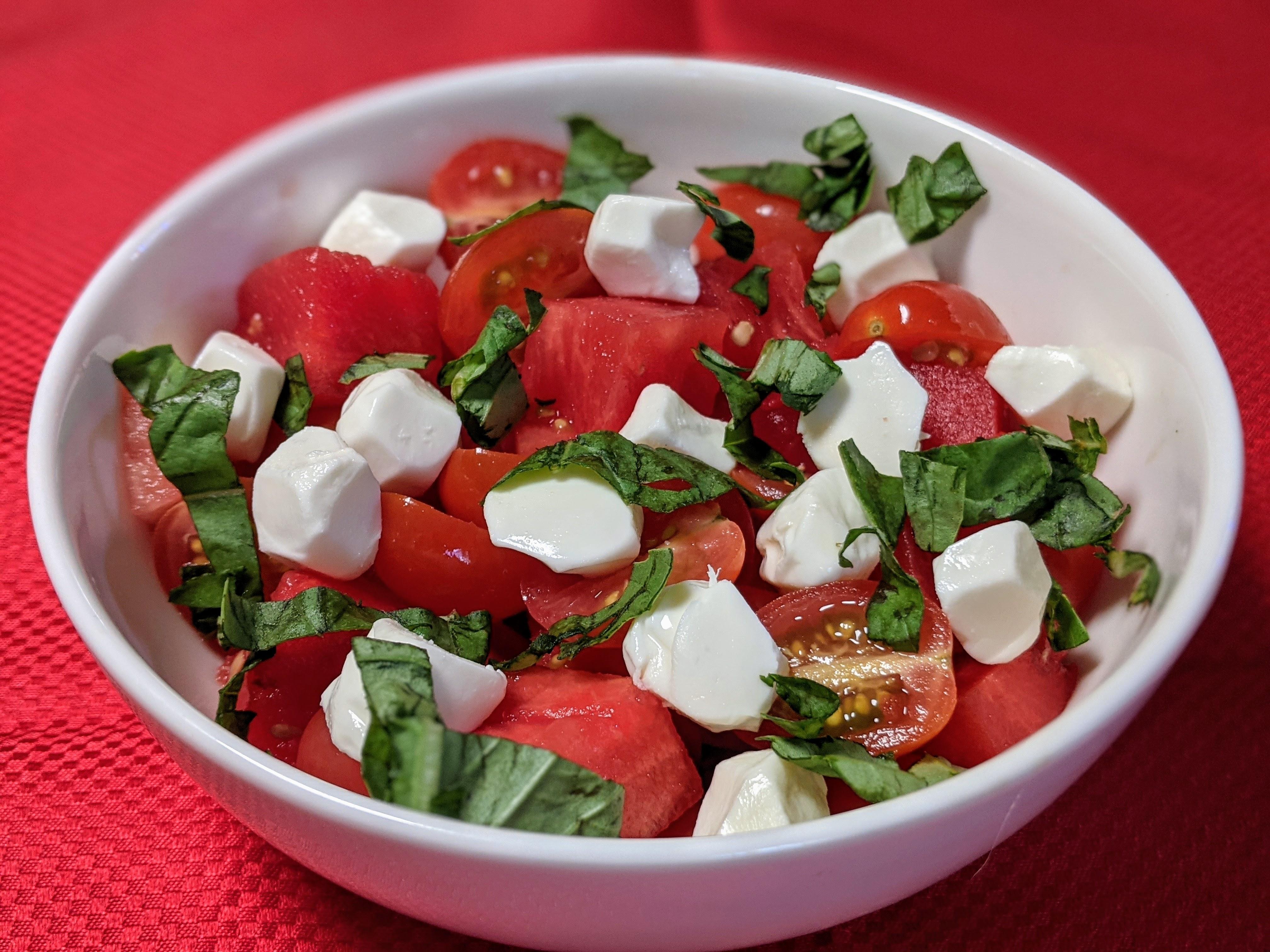 Watermelon, tomato and mozzarella cheese in a white round bowl with a red background.