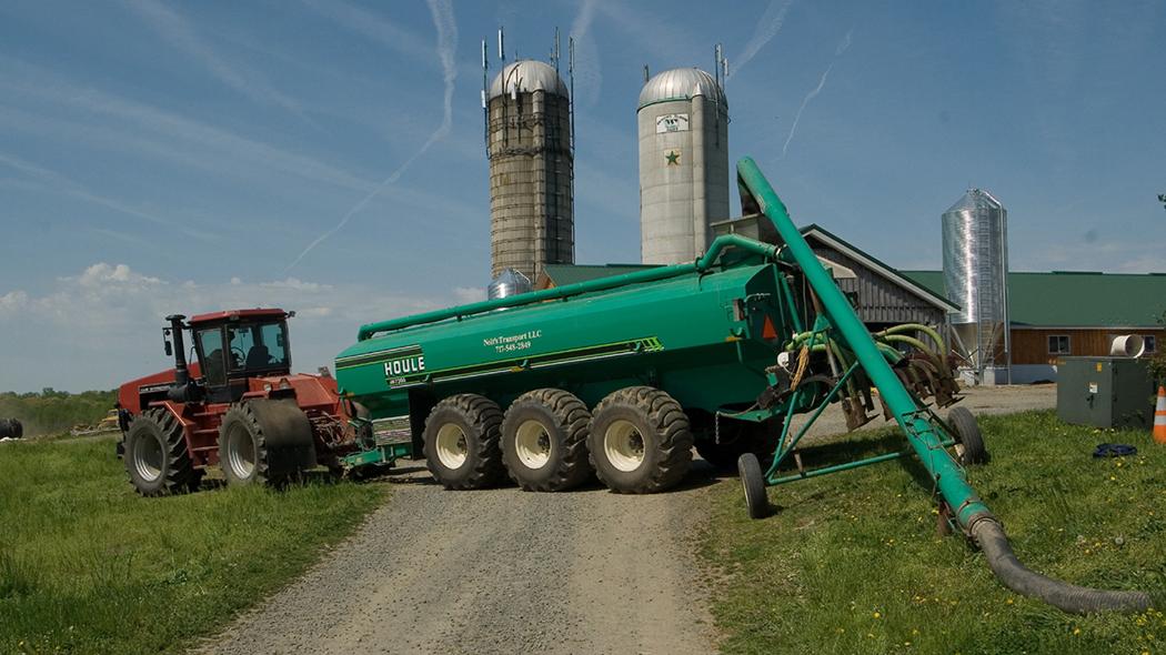  On a gravel driveway in a farmyard, a tractor is hitched to a large green manure injector with multiple wheels and a hose attachment for applying manure or slurry to fields. In the background, there are tall silos and a barn, along with metallic cylindrical storage units.