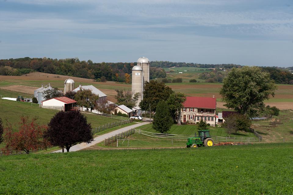 A tractor traverses a field, with a traditional farm house, barn and silos in the background, showcasing rural agricultural life.