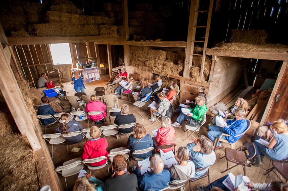 A group of people seated in chairs inside a rustic barn, engaged in conversation or discussion.