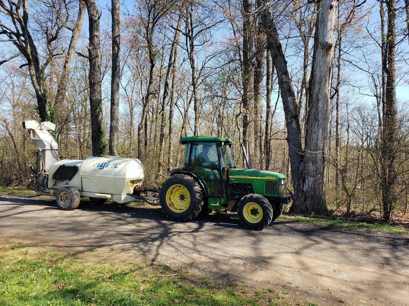 A tractor towing a spray tank along a rural road surrounded by greenery.  