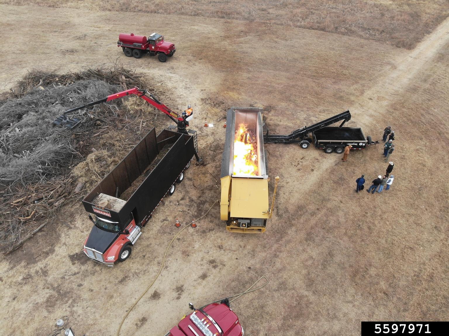 On-site woody biochar production. The woody debris is burned in an air curtain burner (center). The resulting biochar is extinguished and piled in a collection trailer (right ). Photo by Ryan Armbrust, Kansas Forest Service, Bugwood.org.