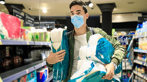 A man wearing a mask and holding several packages of toilet paper in a supermarket.