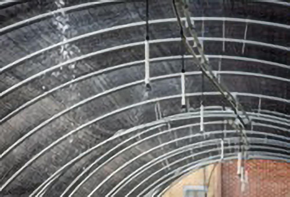 This image shows water being sprayed out of a system of tubes attached to the top of a hoop house with a black cover.