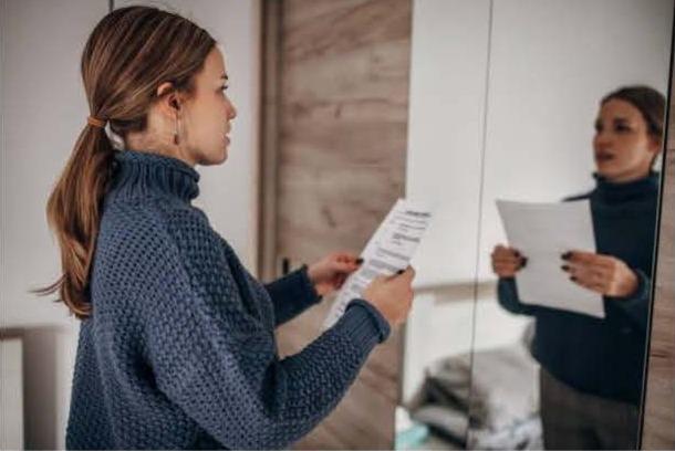 A woman in a blue sweater rehearses lines from a script while facing a mirror. Her focused expression suggests concentration and preparation.
