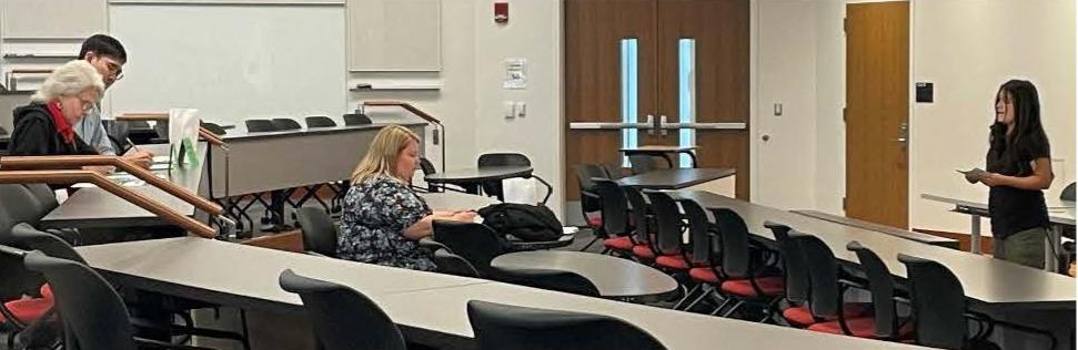 A woman stands at the front of a nearly empty lecture hall, holding papers. Three people sit at desks, listening attentively.