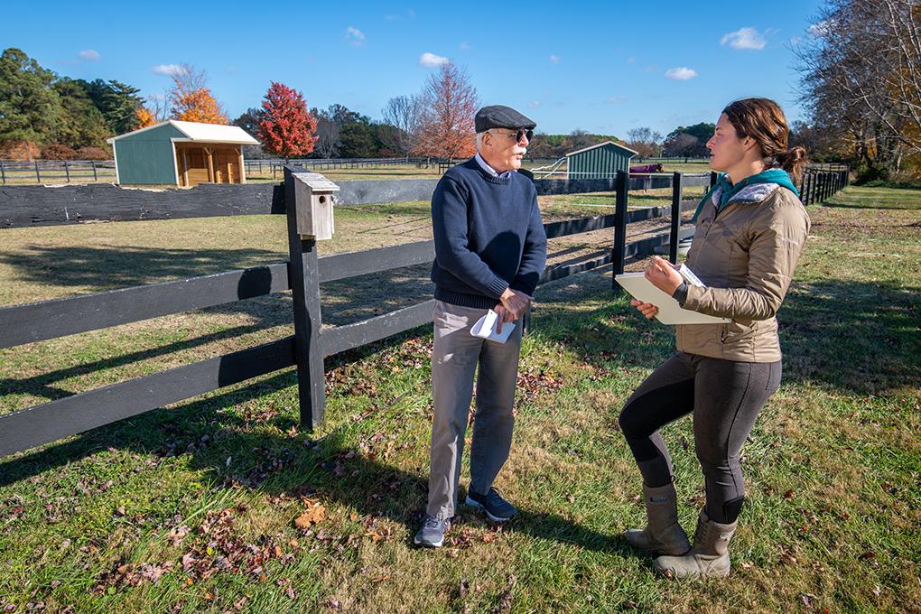 A man in a dark sweater and cap talks to a woman holding a clipboard on a sunny farm. They're by a wooden fence with barns and autumn trees nearby.
