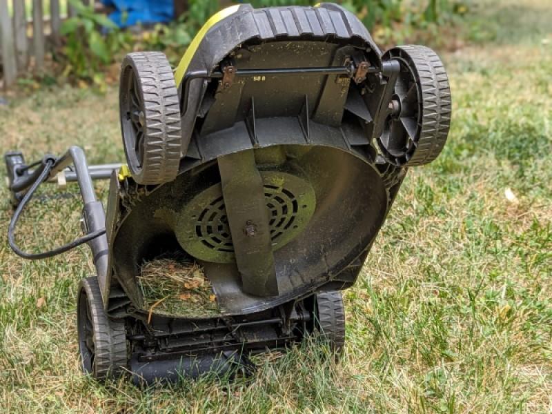 Electric lawn mower propped up on its rear wheels to expose the mower blade housing underneath. A wad of grass clippings sits in the undercarriage.