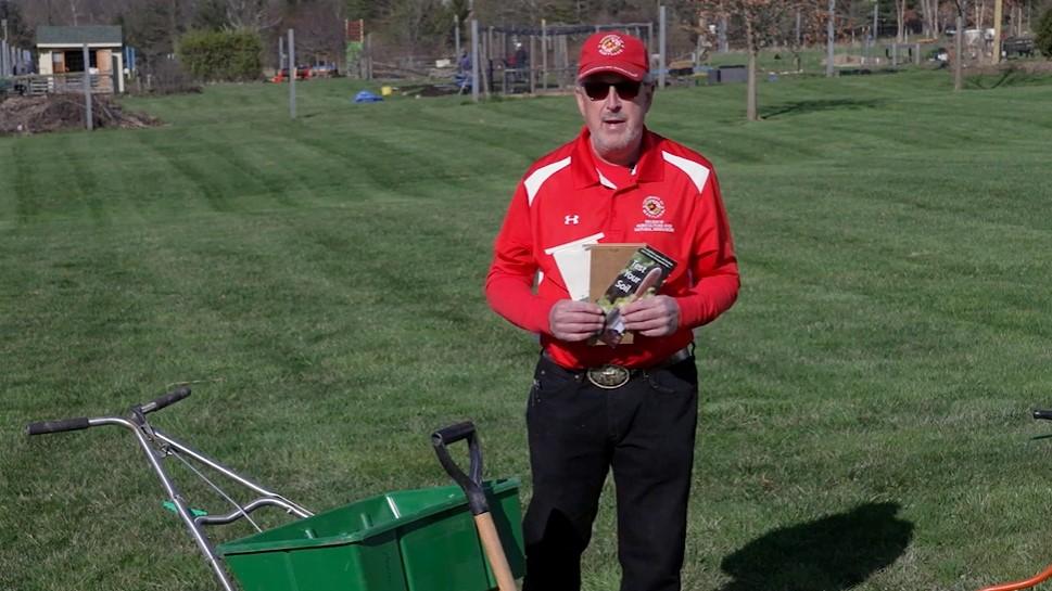 Still image from a video presentation about how to fertilize a home lawn. A presenter is standing in a large lawn behind commonly-used lawn equipment.