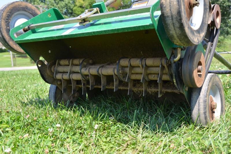 The front of a dethatcher machine raised up to view the underside, which contains a row of multiple metal hoe-like blades arranged vertically to cut grooves into the soil.