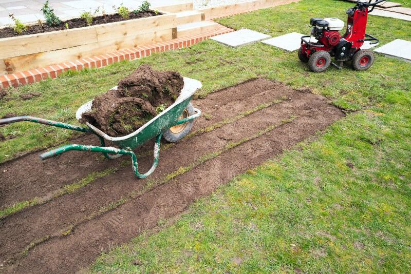 A sod cutter machine sits next to a patch of lawn that has had the sod removed and stacked into a wheelbarrow. The ground where sod was removed is bare soil.