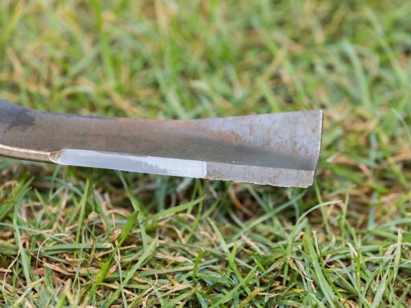 Close-up of a lawn mower blade, removed from the mower. Half of one of the cutting edges has been sharpened and looks clean and polished.