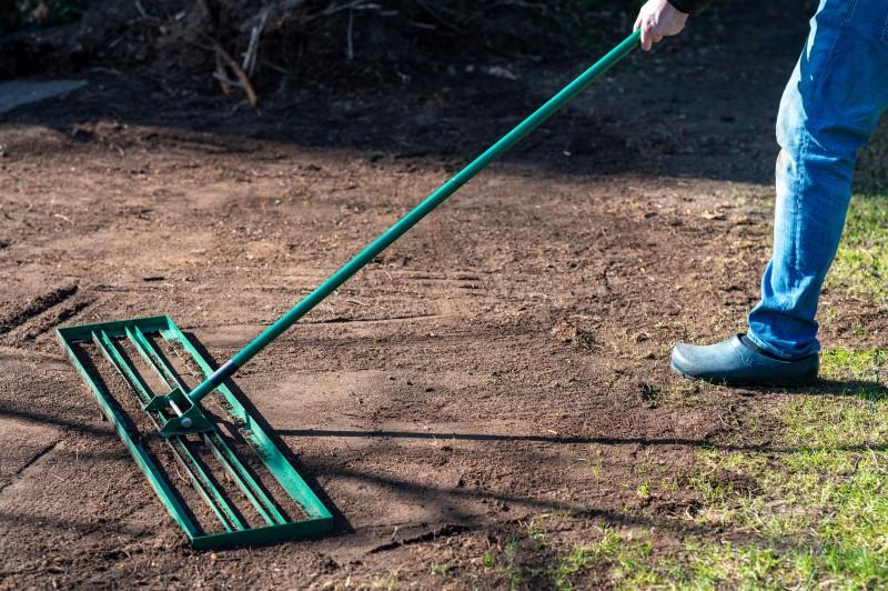 A person using a wide, flat, slotted metal rake to level-out bare soil.
