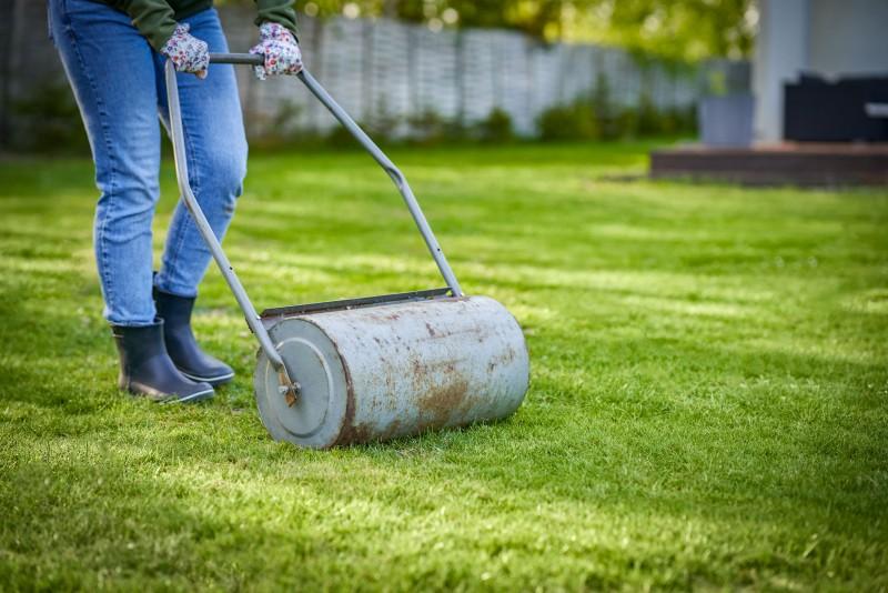 A person uses a handle to roll a metal cylinder over a lawn.