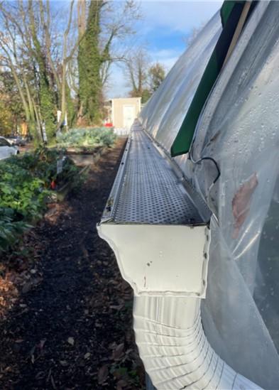 A close‑up view of a metal gutter attached along the side of a plastic‑covered greenhouse. The gutter leads into a white downspout that curves downward. Raised garden beds, leafy plants, and tall trees line the path beside the greenhouse under a bright sky.