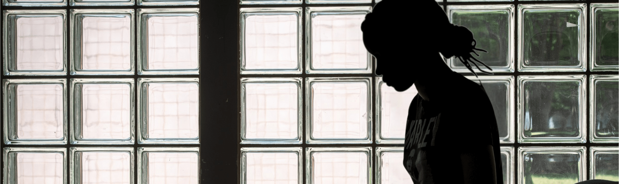 Dark, high-contrast silhouette of a person in side profile standing near a wall of square glass block windows. The person’s face and upper body are blurred and heavily shadowed, making individual features indistinguishable, with only a softened outline and hair shape visible against the softly lit background.