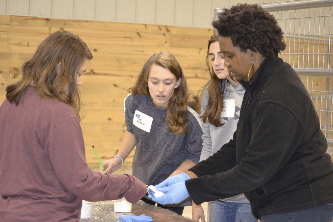 Four people gathered around a table indoors, participating in a hands-on activity with small containers. One person wearing gloves demonstrates or assists while the others observe and handle materials. The setting includes a wooden wall and a metal enclosure in the background.
