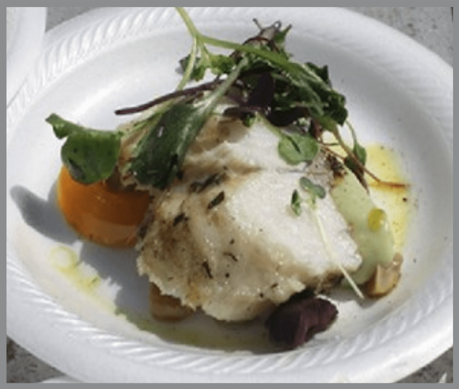 A plated dish featuring a white fish fillet topped with herbs and greens, served on a ridged white plate with a light sauce. The fish rests on a bed of vegetables, including a visible piece of orange-colored squash, with small garnishes and greens arranged on top.