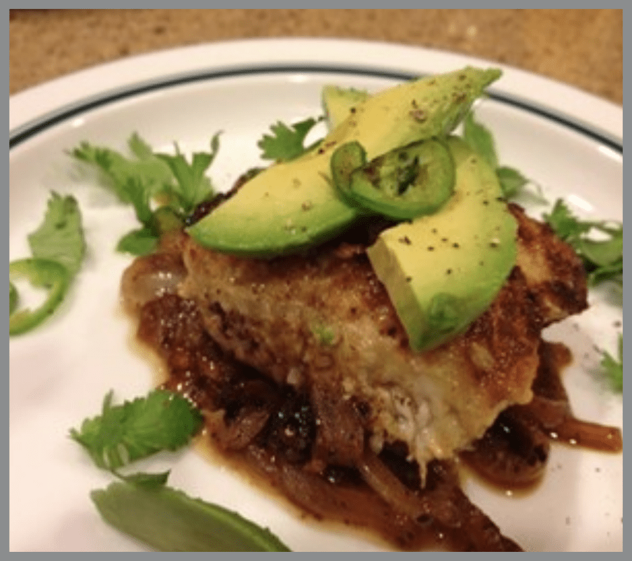 Un plato servido que presenta un filete de pescado de color marrón, bien sazonado, cubierto con aguacate y cilantro, servido en un plato blanco acanalado.
