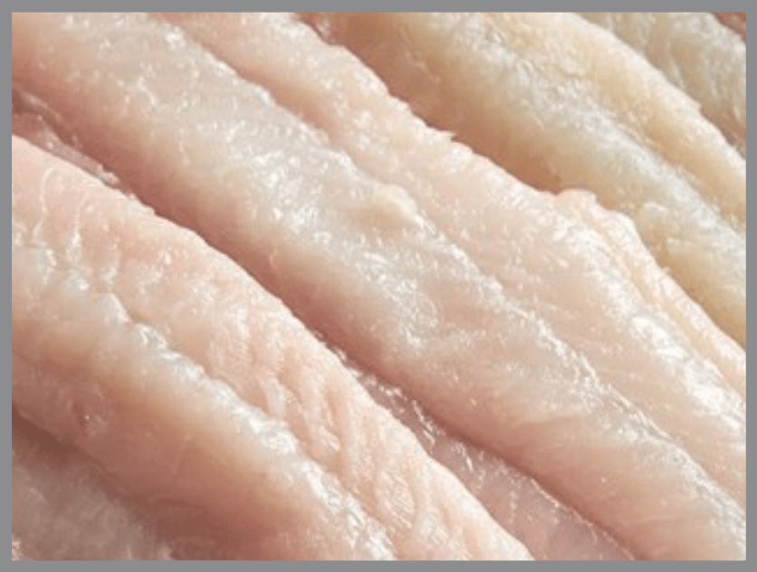 Vista de cerca de varios filetes de pescado crudos de color rosado pálido, dispuestos uno junto al otro, que muestran superficies lisas y ligeramente brillantes, con surcos visibles y una textura fina que recorre su longitud.