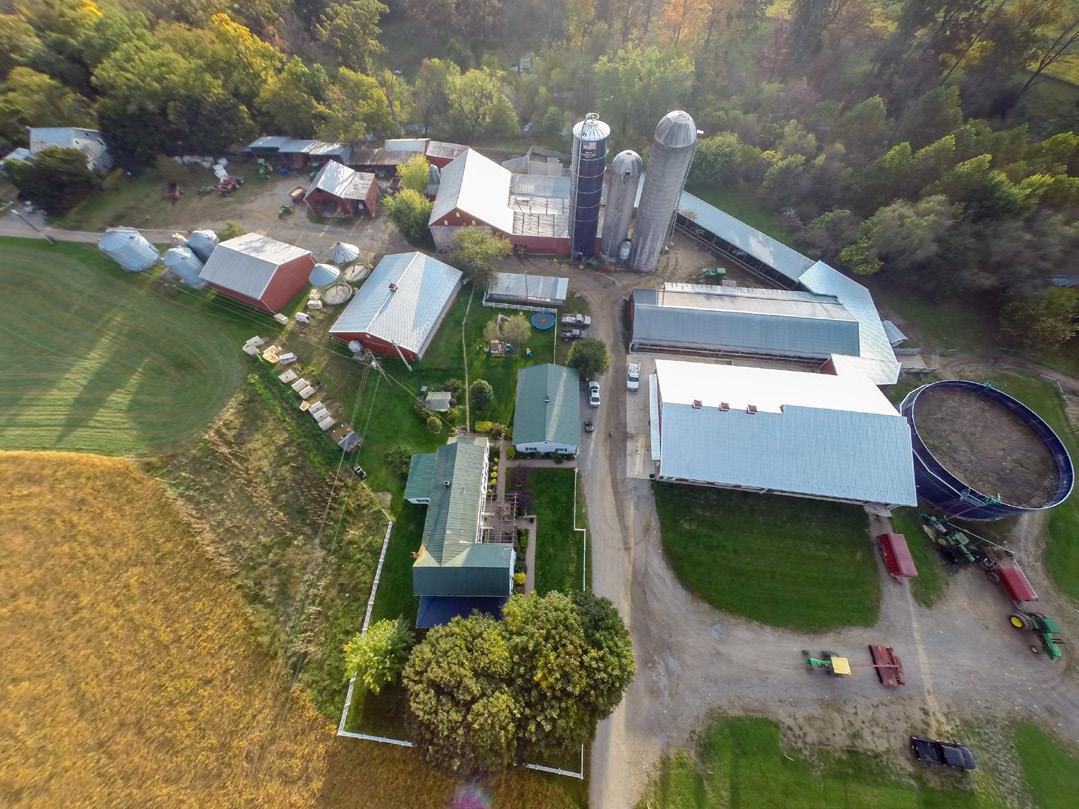 Overhead view of a farm, showing the roofs of multiple barns, a home, small silos and a manure holding tank. Various tractors are driving around, and the farm fields are a lush green and gold.