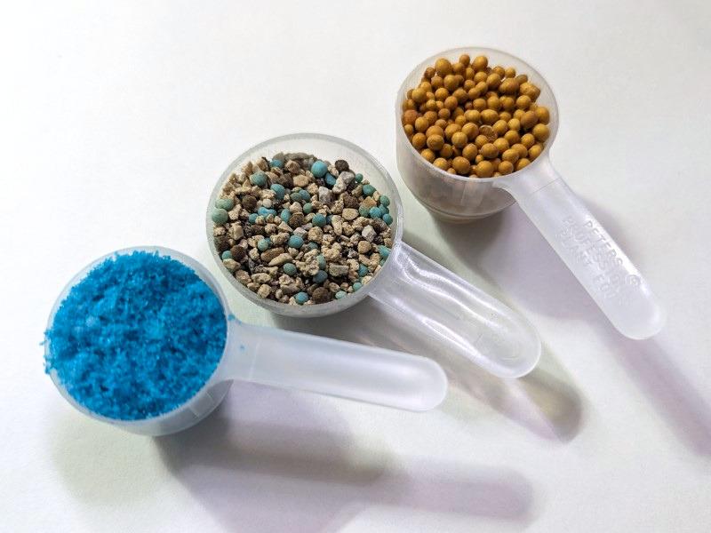 Three similarly-sized plastic scoops, each holding a different fertilizer. One is dyed blue and somewhat powdery; one consists of three colors of pellets; one has round pellets of uniform color and size.