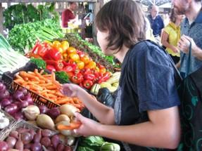 Person selecting fresh produce at an outdoor market, standing beside a display of colorful vegetables including peppers, carrots, onions, potatoes, and leafy greens arranged in baskets and crates.